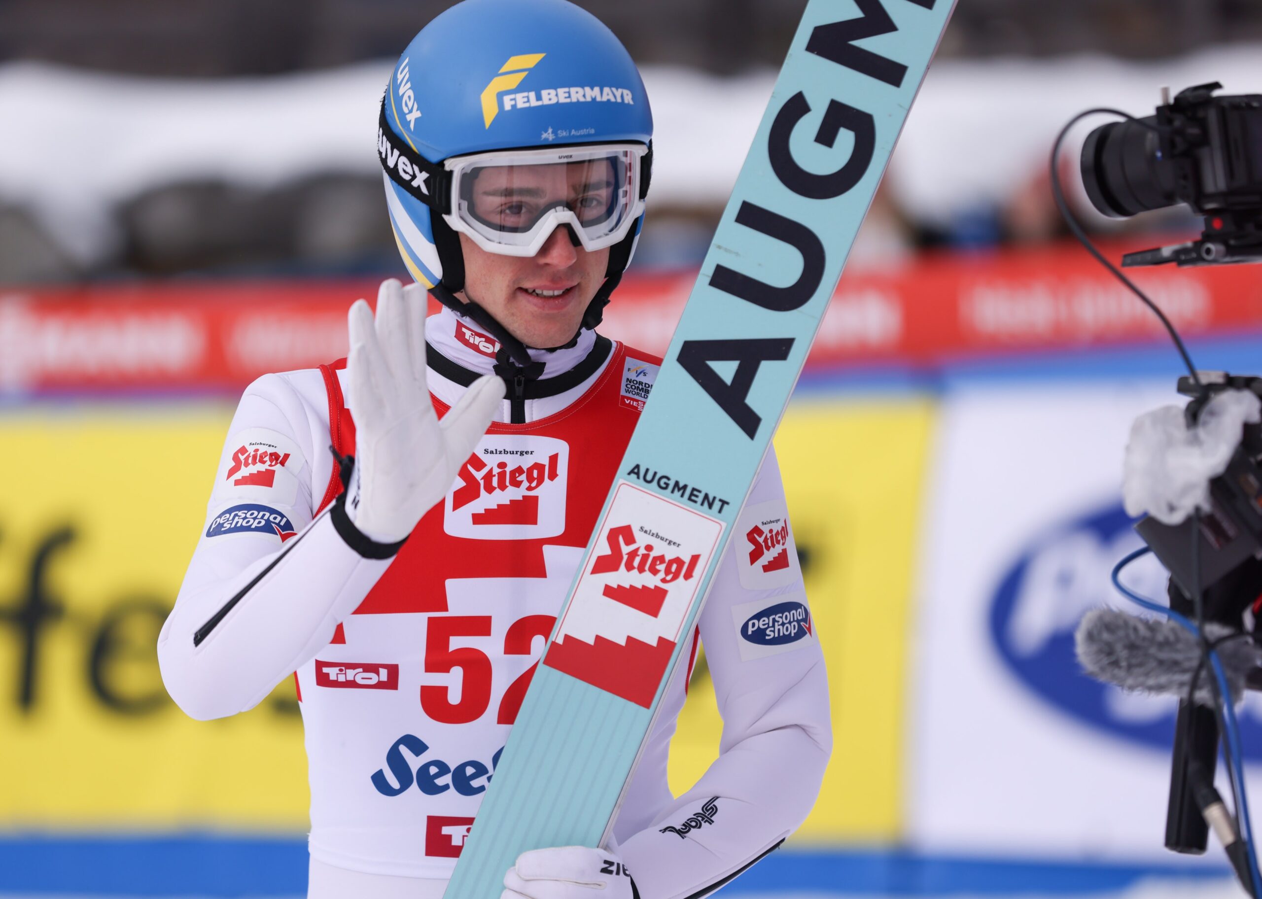 Ein Skispringer in einem weiß-blauen Trikot winkt mit seinen Skiern in der Hand in die Kamera.