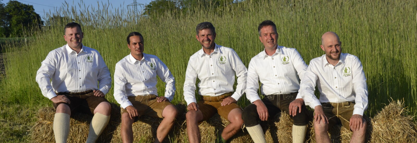 Fünf Männer in weißen Hemden und traditionellen österreichischen Lederhosen sitzen auf einem Heuballen auf einem Feld.