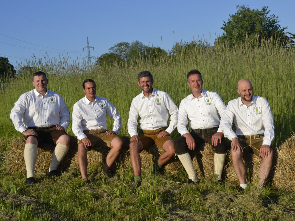 Fünf Männer in weißen Hemden und traditionellen österreichischen Lederhosen sitzen auf einem Heuballen auf einem Feld.