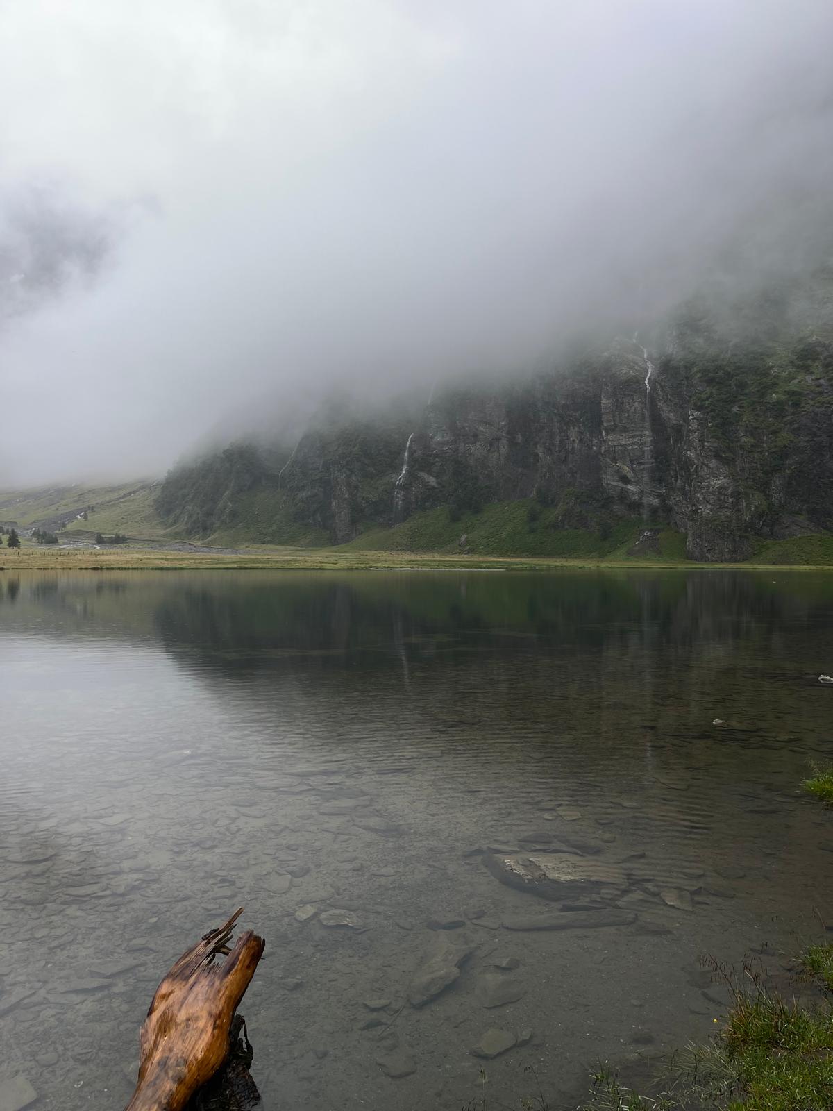 Ein ruhiger See, in dem sich ein nebelverhüllter Berg mit Wasserfällen spiegelt.