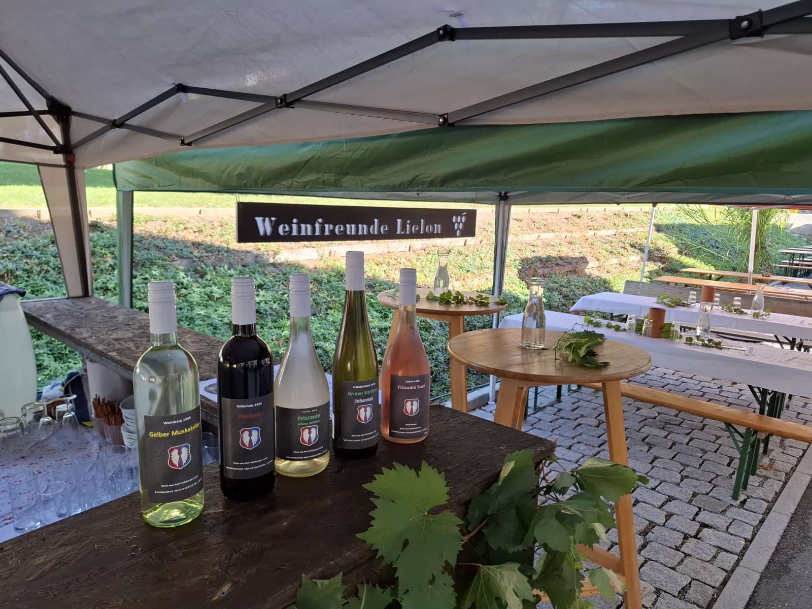 Fünf Weinflaschen mit Etiketten von Weinfreunde Lielon, ausgestellt auf einer hölzernen Außenbar.