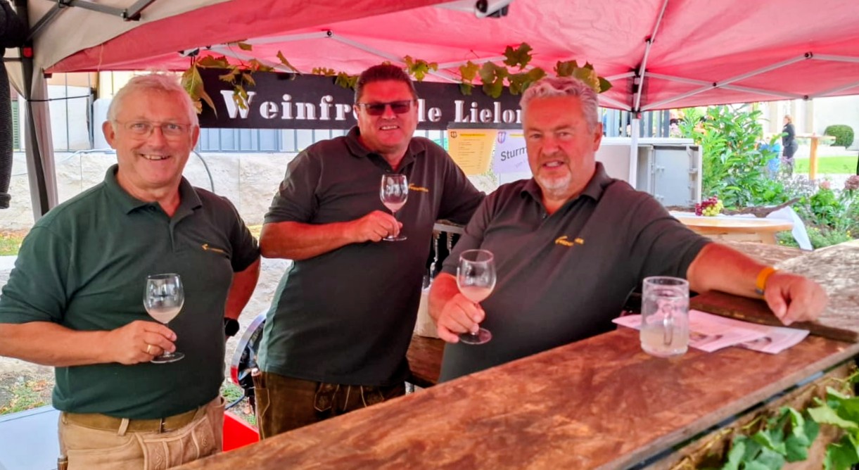 Drei Männer in olivfarbenen Poloshirts und Lederhosen stehen hinter einer Bar bei einer österreichischen Veranstaltung im Freien; jeder hält ein Glas Wein in der Hand.