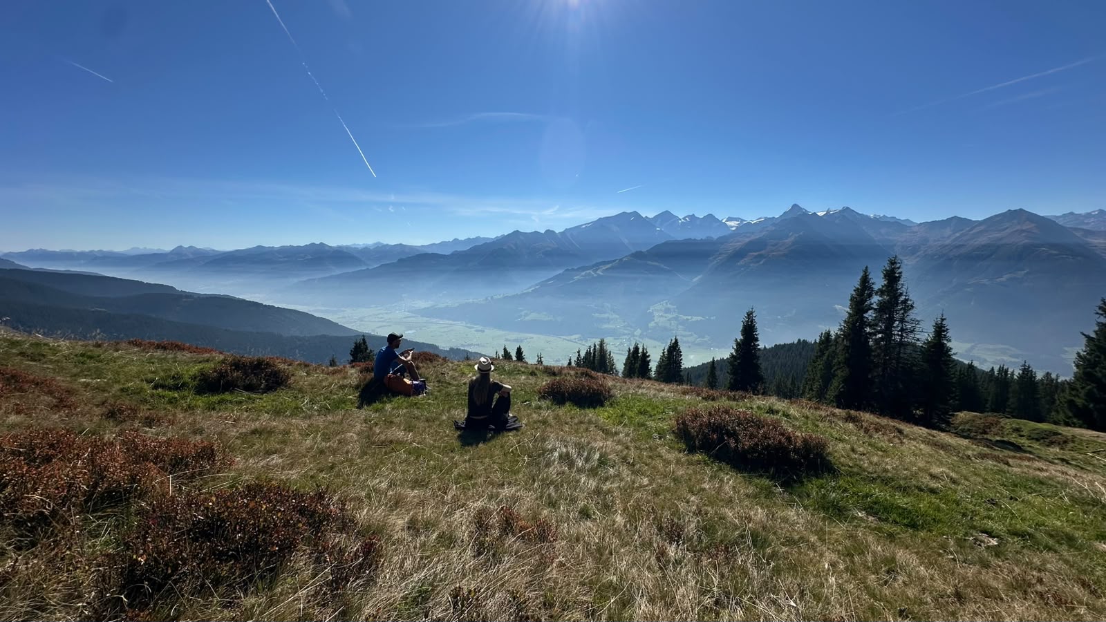 Zwei Menschen sitzen auf einem grasbewachsenen Berghang im Herbst und blicken über ein nebliges Tal in Richtung der österreichischen Alpen.