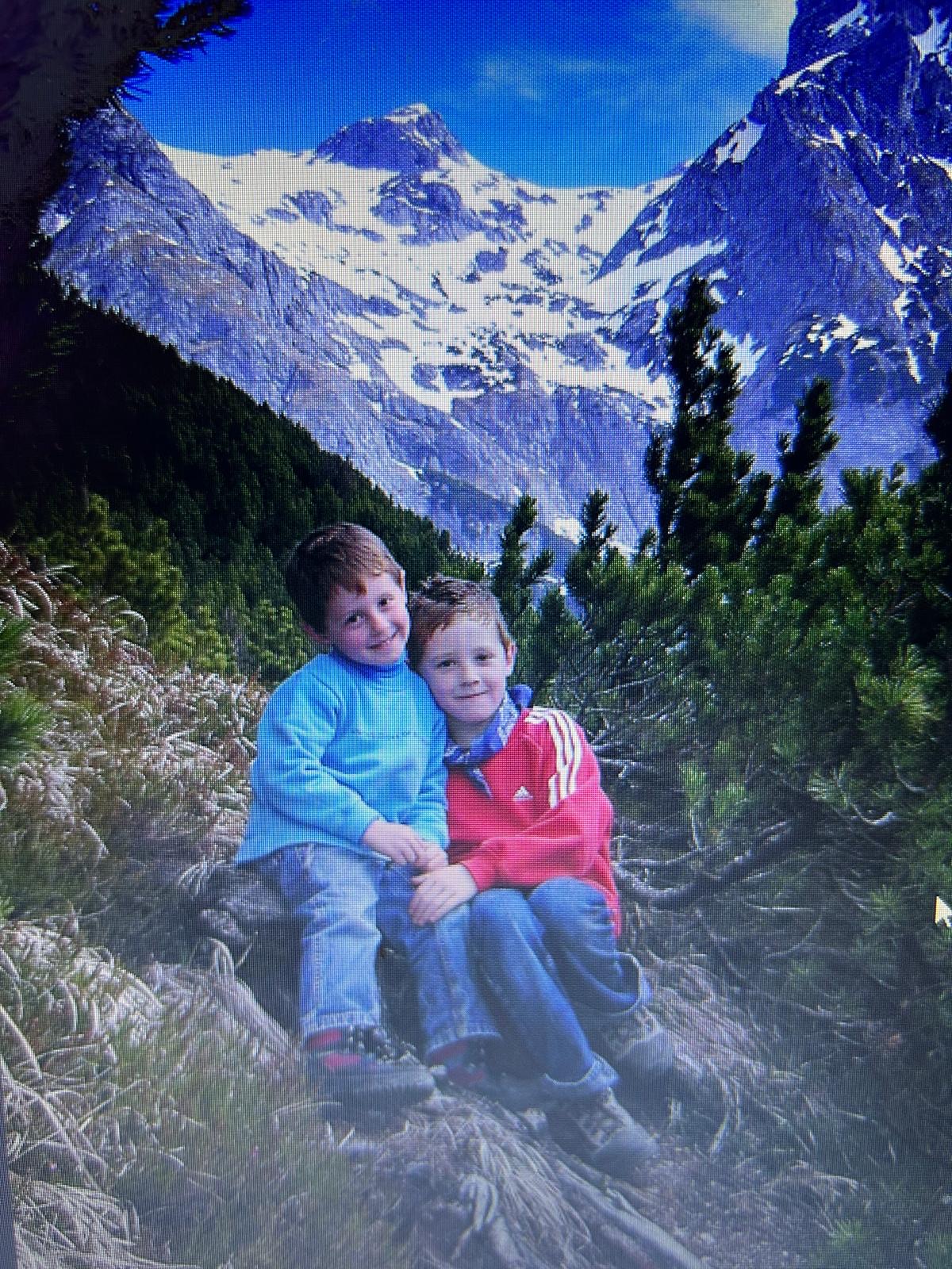 Zwei kleine Jungen sitzen zusammen an einem Berghang vor einer schneebedeckten österreichischen Bergkette.