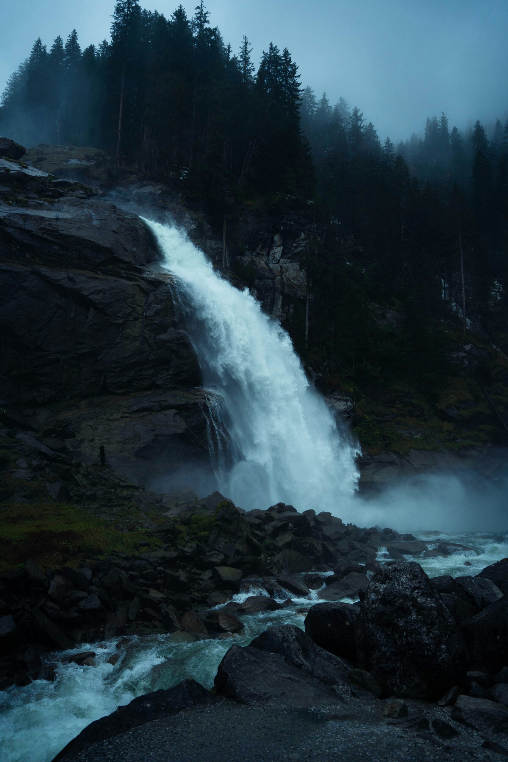 Ein großer Wasserfall stürzt über felsige Klippen in einen Bach hinab, umgeben von einem dunklen, nebligen Kiefernwald.