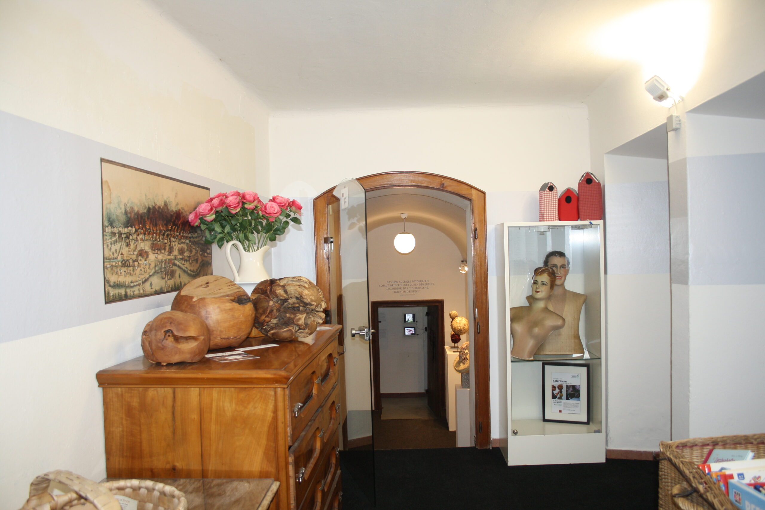 Eine Kommode aus Holz mit Schalen und Rosen, ein gewölbter Türdurchgang und eine Vitrine mit zwei Mannequin-Büsten.