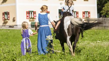 Ein Kind reitet auf einem Pony, die Mutter und das andere Kinder gehen händchenhaltend nebenbei her (Rückansicht)